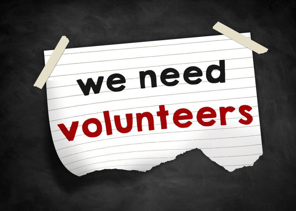A lined white paper is taped to a blackboard. It states, "we need volunteers"